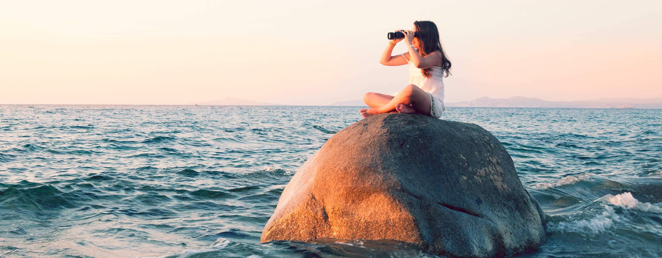 Mädchen sitzt mit Fernglas auf einem Felsen im Meer.
