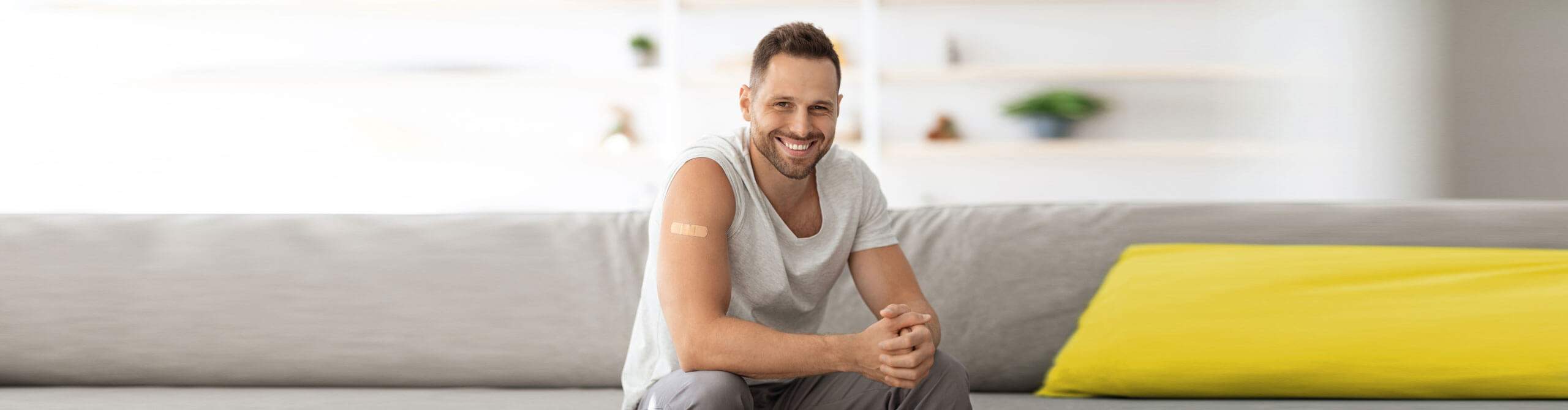 Ein junger Mann mit einem Pflaster auf dem Oberarm sitzt lächelnd auf einem Sofa