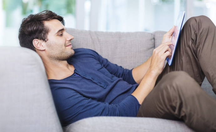Mann schaut auf dem Sofa liegend lächelnd auf ein Tablet.e